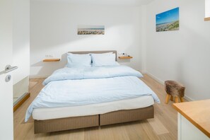 A bedroom with a bed, bedside tables, a wooden stool, and two wall-mounted artworks. - Modern vacation apartment for 4 people, new building near the beach (Juist)
