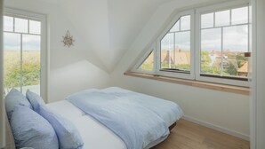 A bedroom with a sloped ceiling, a bed with blue bedding, a window with a view of houses, and a wall-mounted decoration. - Modern vacation apartment for 4 people, new building near the beach (Juist)