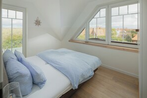 A bedroom with a sloped ceiling, a bed with blue bedding, a window with a view of houses, and a wall-mounted decoration. - Modern vacation apartment for 4 people, new building near the beach (Juist)