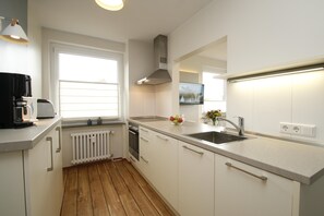A modern kitchen with white cabinets, a stainless steel sink, and a window with blinds. - "Experience pure relaxation in the charming vacation apartment on Juist!" (Juist)