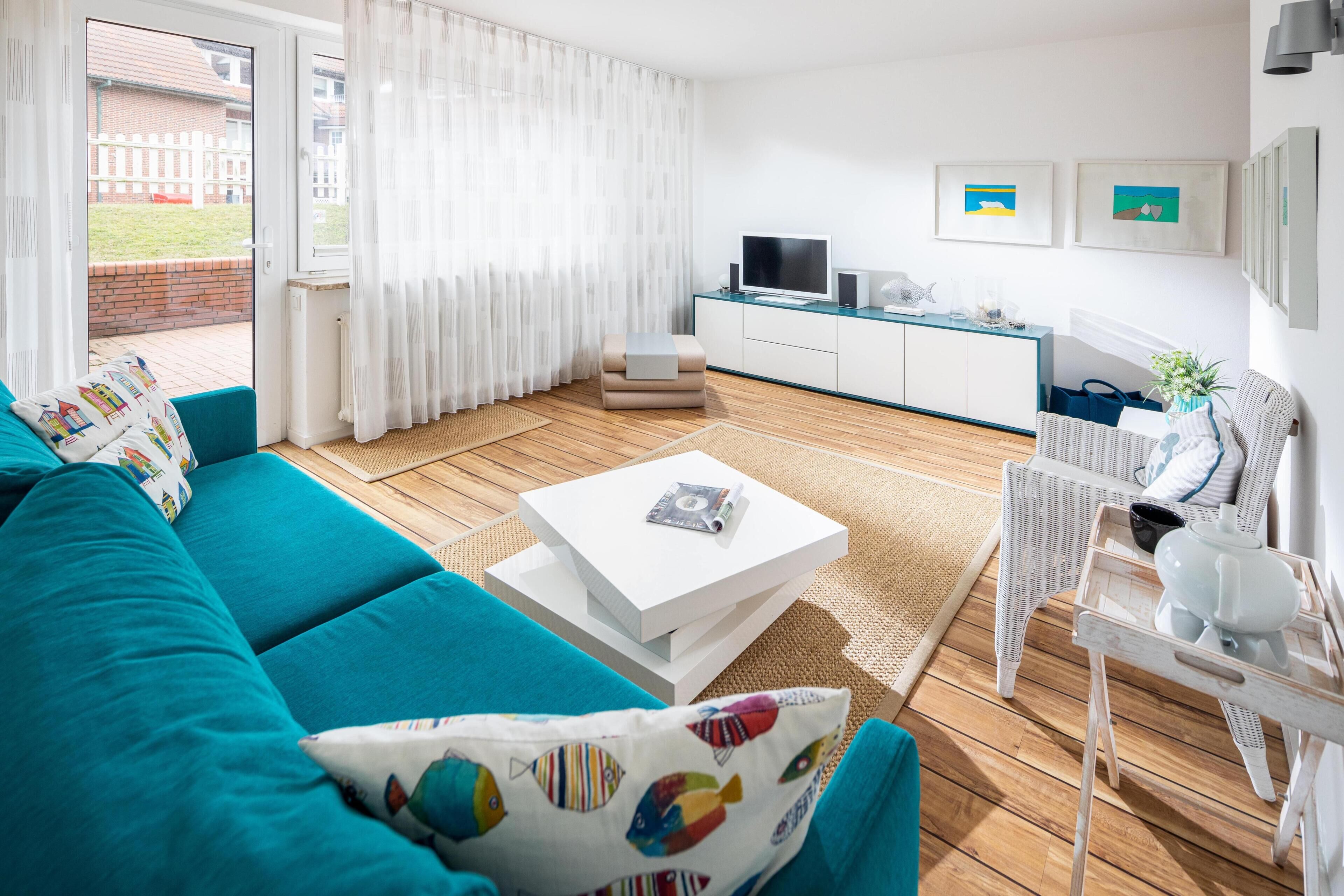 A modern living room with a teal sofa, a white coffee table, and a kitchen area with a TV and framed pictures.