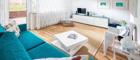 A modern living room with a teal sofa, a white coffee table, and a kitchen area with a TV and framed pictures.