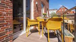 Een balkon met gele rieten stoelen en een tafel, een glazen deur en een bakstenen gebouw op de achtergrond.