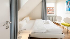 A modern hotel room with a bed, a desk, a chair, and a window with a view of a building. - Centrally located vacation apartment for 3 people (Juist)