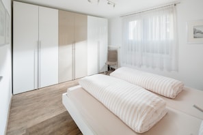 A bedroom with a bed, a wardrobe, a chair, and a window with curtains. - Cozy vacation apartment with garden and close to the beach (Juist)