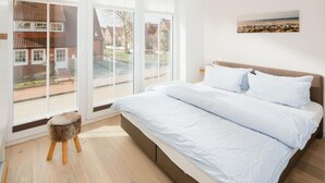 Ett sovrum med en stor säng, en liten pall och utsikt över ett bostadsområde genom stora fönster.