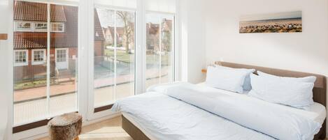 A bedroom with a large bed, a small stool, and a view of a residential area through large windows.