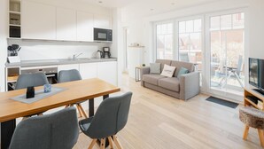 A modern kitchen with white cabinets, a microwave, and a coffee maker. There is a dining table with chairs, a grey sofa with a cushion, and a television on a wooden stand. - Modern vacation apartment for 2 persons, new building near the beach (Juist)