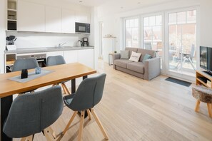 A modern kitchen with white cabinets, a microwave, and a coffee maker. There is a dining table with chairs, a grey sofa with a cushion, and a television on a wooden stand. - Modern vacation apartment for 2 persons, new building near the beach (Juist)