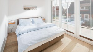 A modern bedroom with a bed, wooden flooring, a bedside table, and a window with a view of buildings. - Modern vacation apartment for 2 persons, new building near the beach (Juist)