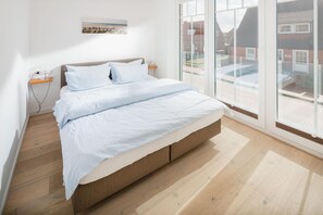 A modern bedroom with a bed, wooden flooring, a bedside table, and a window with a view of buildings. - Modern vacation apartment for 2 persons, new building near the beach (Juist)