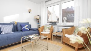 Uma sala de estar com um sofá azul, cadeiras de vime, uma mesa de centro de vidro e vista para um veleiro pela janela.