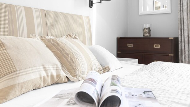 Uma cama bem arrumada com uma revista, um travesseiro xadrez e uma mesa de cabeceira de madeira com um recipiente de vidro.