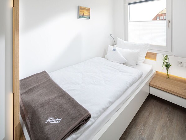A single bed with a white mattress and pillows, a brown blanket, a small bedside table with a plant, and a window with blinds.