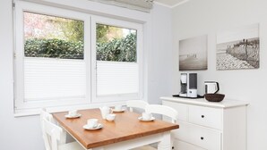 Une pièce avec une table en bois, des chaises blanches, un meuble blanc et deux fenêtres équipées de stores.