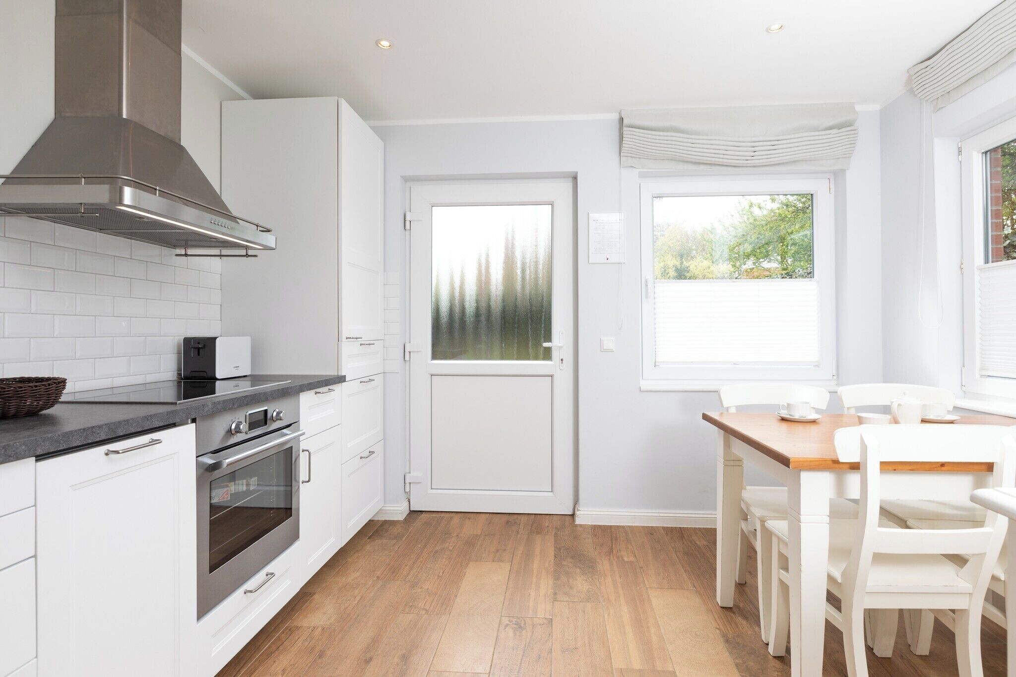 Una cocina moderna con armarios blancos, una campana extractora de acero inoxidable, horno empotrado y un comedor con mesa de madera y sillas blancas.