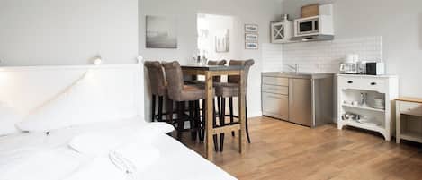 Quarto com uma cama grande, uma mesa de jantar de madeira com cadeiras, uma cozinha com armários brancos e um micro-ondas.