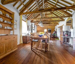 Una habitació àmplia amb bigues de fusta, una taula de menjador amb caires i una zona de cuina amb armaris de fusta i una estufa.