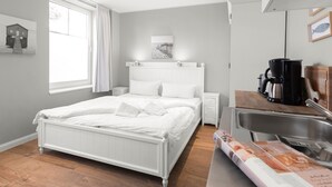 Un dormitorio ordenado con cama, mesitas de noche, ventana con cortinas y una pequeña cocina con lavamanos y cafetera.