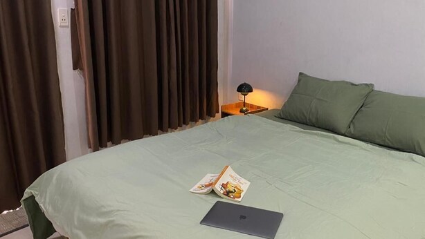 Een netjes opgemaakt bed met een laptop en een boek erop, een bedlampje en een raam met gordijnen.