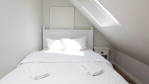 Un dormitorio con techo inclinado, una cama con ropa de cama blanca, una mesita de noche y un ventanal.