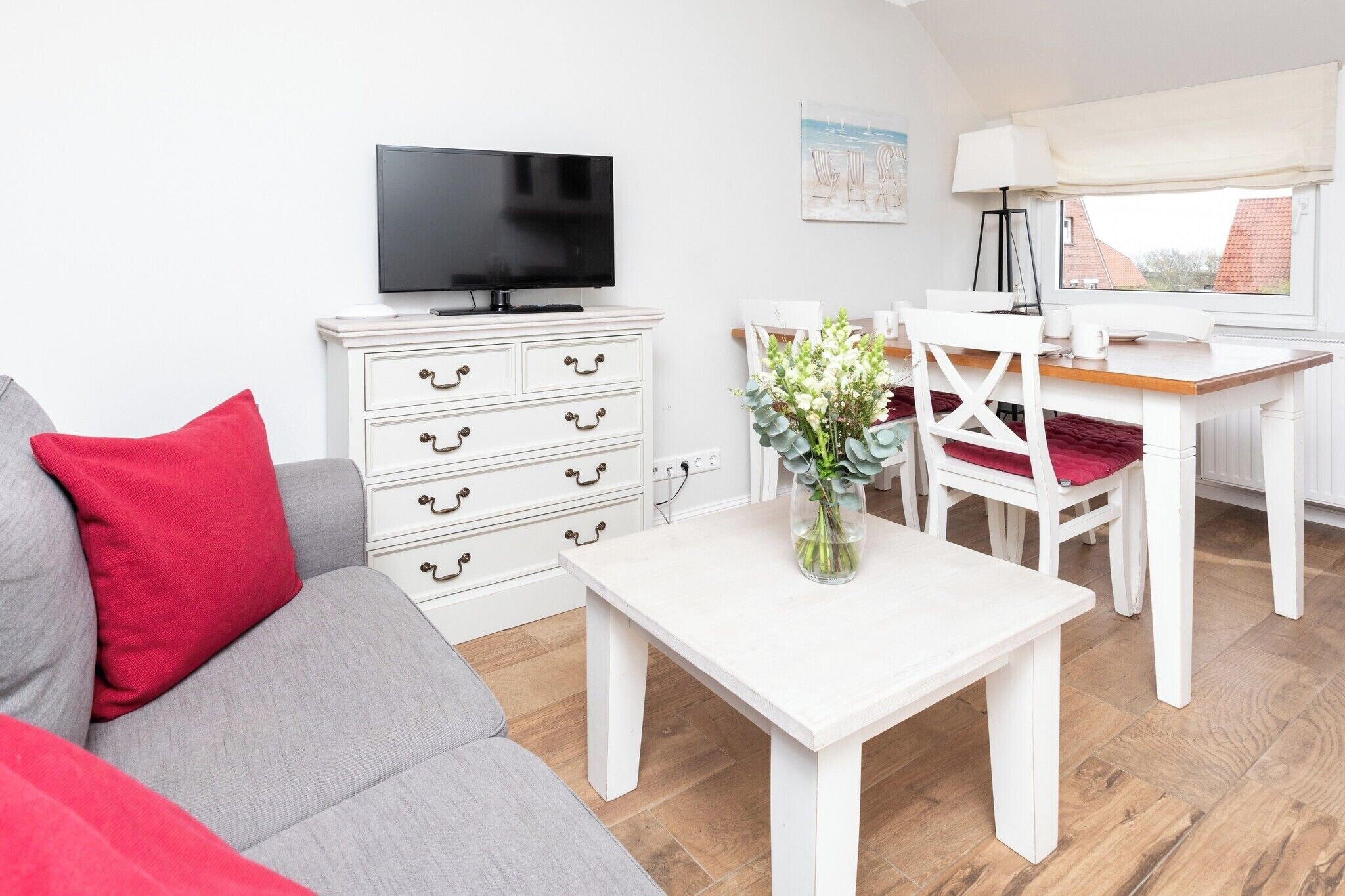 Un salon comprenant un canapé, un téléviseur, une table blanche avec un vase, et un coin repas avec des chaises et une table.