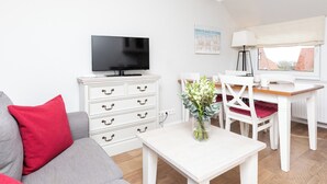 Una sala de estar con un sofá, un televisor, una mesa blanca con un florero y un comedor con sillas y una mesa.
