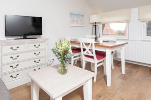 A room with a TV on a dresser, a dining table with chairs, a vase with flowers, and a window with a view. - Apartment in a quiet location with balcony (Juist)