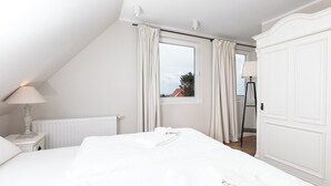 Un dormitorio blanco con cama, mesita de noche con lámpara, ventana con cortinas y un radiador.
