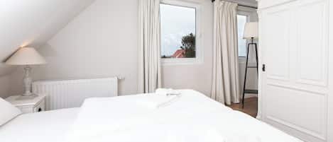A white bedroom with a bed, a nightstand with a lamp, a window with curtains, and a radiator.