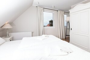 A white bedroom with a bed, a nightstand with a lamp, a window with curtains, and a radiator. - Apartment in a quiet location with balcony (Juist)