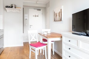 A room with a wooden dining table, white chairs, a white TV stand, and a door leading to another room.