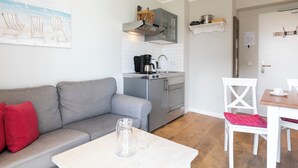 Ein kompakter Wohnbereich mit einer Küchenzeile, einem grauen Sofa mit roten Kissen, einem Esstisch mit einem weißen Stuhl und einem Bild von Strandstühlen an der Wand.