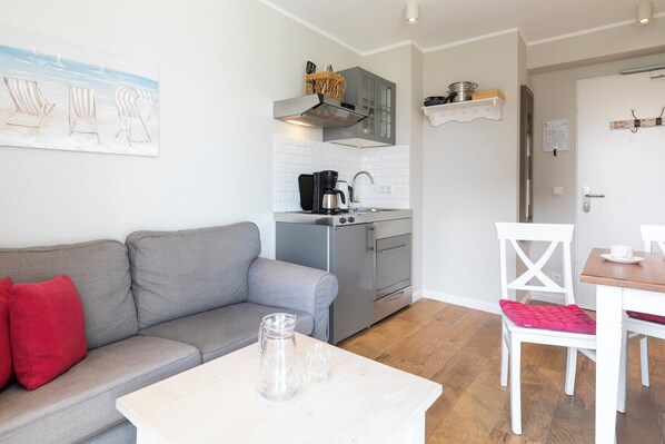 A compact living space with a kitchenette, a grey sofa with red cushions, a dining table with a white chair, and a painting of beach chairs on the wall.