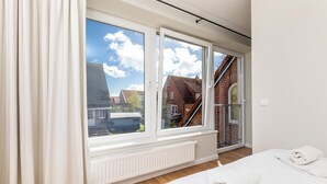 Ein Schlafzimmer mit großem Fenster, Heizkörper, einem Bett mit weißer Bettwäsche und Blick auf ein Backsteinhaus.
