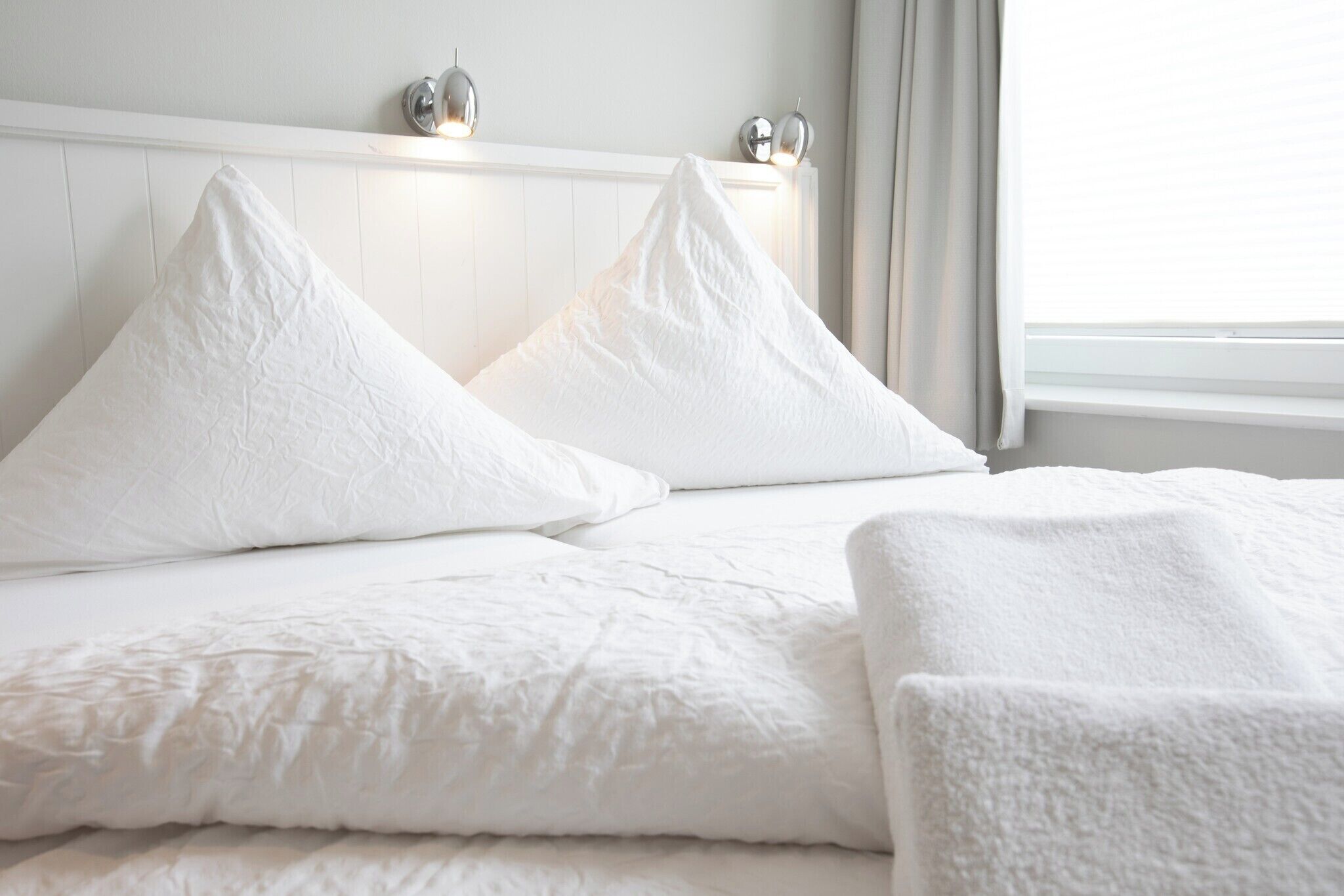 A bed with white bedding and pillows, a white headboard, and a window with curtains.