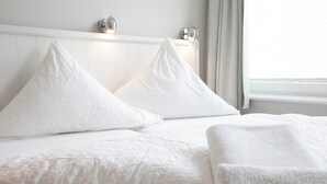 Ein Bett mit weißer Bettwäsche und Kissen, ein weißes Kopfteil und ein Fenster mit Vorhängen.