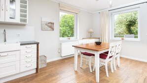 Una cocina moderna con armarios blancos, una mesa de comedor de madera y sillas. Hay ventanas que ofrecen luz natural y vistas a zonas verdes.