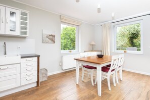 A modern kitchen with white cabinetry, a wooden dining table, and chairs. There are windows providing natural light and views of greenery. - Apartment in a quiet location with balcony (Juist)