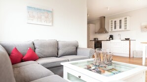 Una sala de estar moderna con un sofá gris, cojines rojos, una mesa de centro de vidrio y una cocina al fondo.