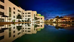 A modern hotel with a pool reflecting the building and palm trees, a covered area with seating, and a clear night sky. - Beach Apartment with Pool, Terrace & Parking (Velilla-Taramay)