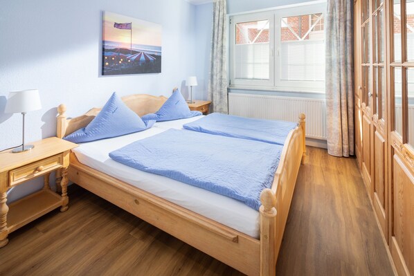 Ein Doppelbett mit Holzrahmen, zwei Nachttische mit Lampen, ein Fenster mit Jalousien und ein Wandbild mit Strandmotiv.