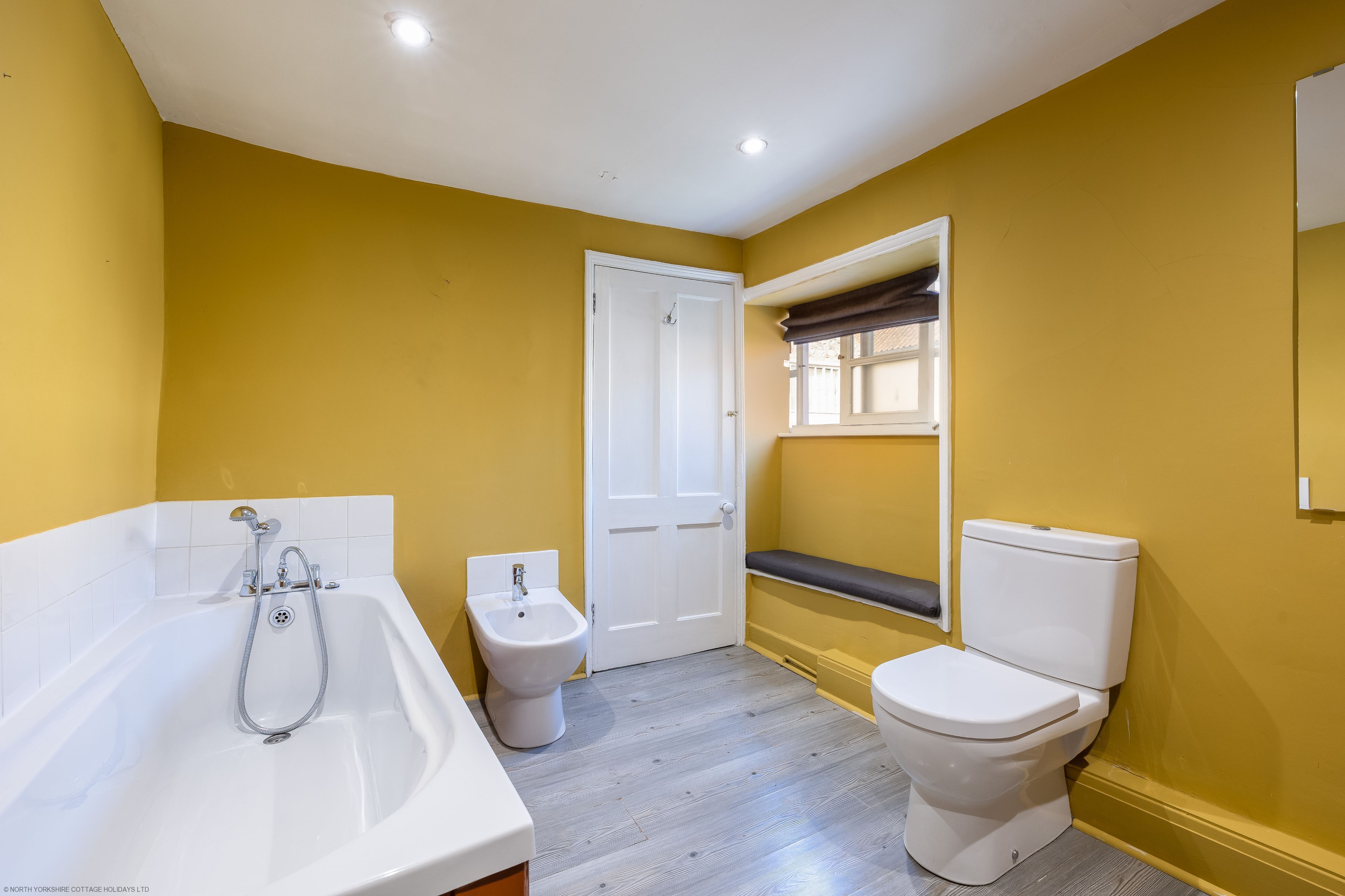 A bathroom with a bathtub, toilet, and bidet, featuring yellow walls and wooden flooring.