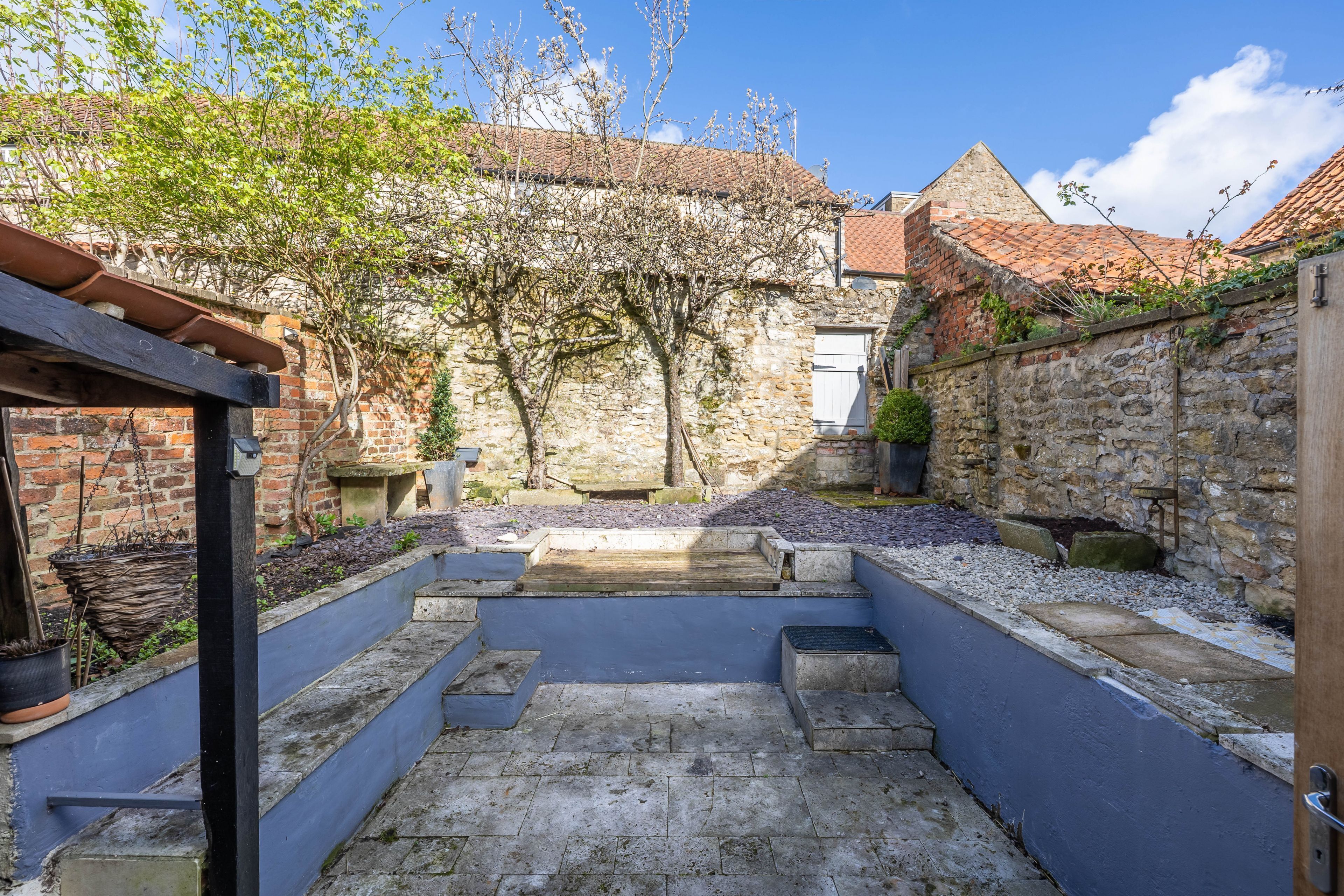 A courtyard with stone walls, a wooden pergola, and a view of a building with a tiled roof.