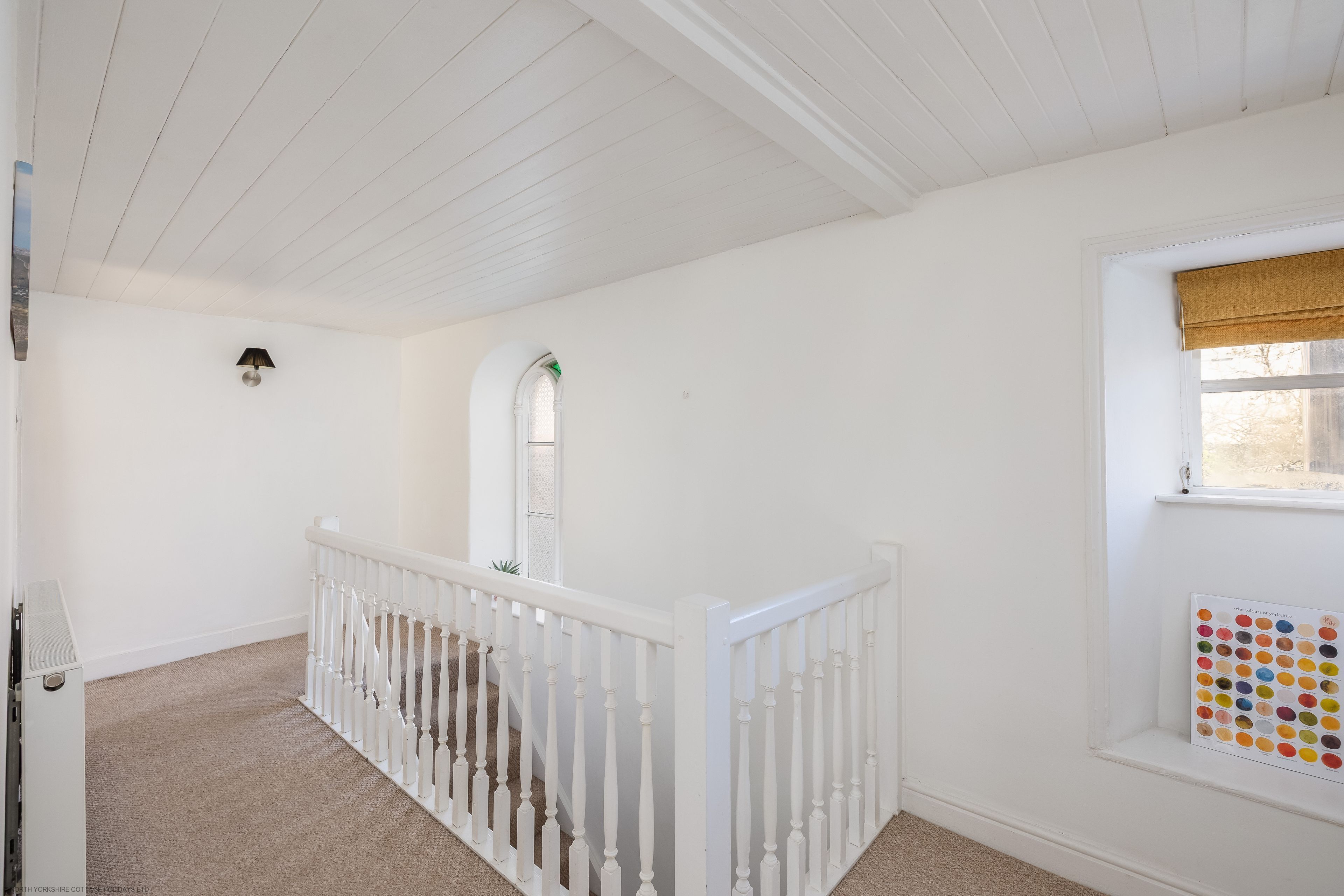 Een kamer met witte wanden, een houten plafond, een witte balustrade, een raam met uitzicht en een aan de muur bevestigde lamp.