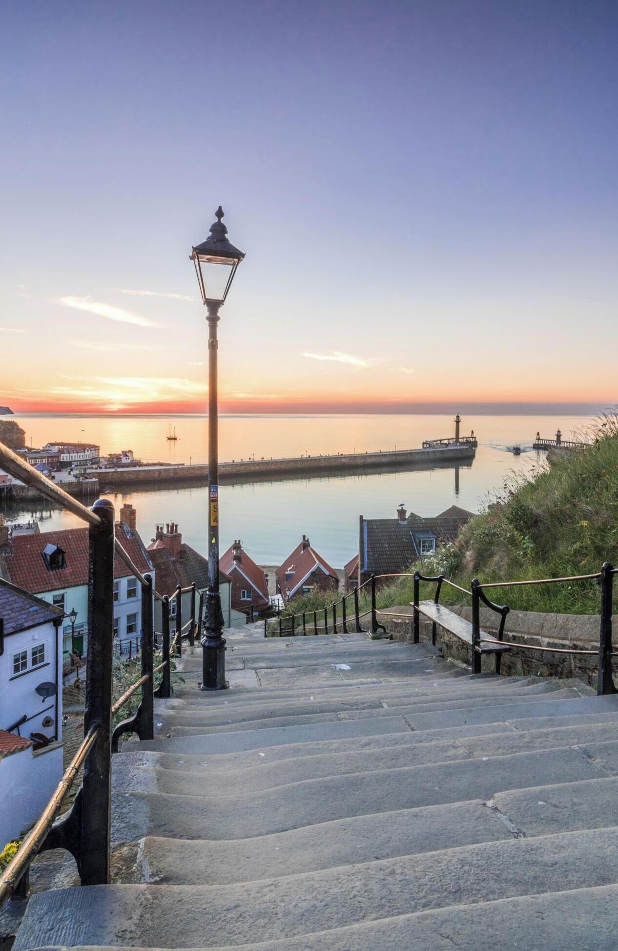 A staircase leading to a waterfront area with a lamp post, stone railings, and buildings with red roofs.