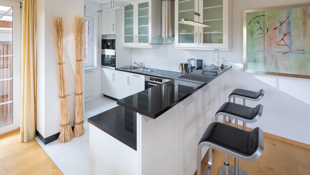 Une cuisine moderne avec des armoires blanches, un comptoir noir et une hotte de cuisinière en acier inoxydable. On y trouve trois tabourets de bar et une grande fenêtre qui laisse entrer la lumière naturelle.