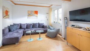 Un salon moderne avec un canapé d’angle gris, une petite table ronde, un pouf gris, un téléviseur et un meuble en bois.