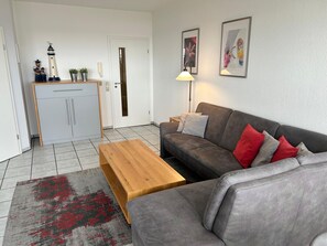 Een woonkamer met een grijze bank, een houten salontafel en een vloerkleed met een rood en zwart patroon.