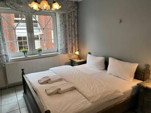 Een netjes opgemaakt bed met witte lakens en handdoeken, een bedlampje, een raam met gordijnen, en uitzicht op een bakstenen gebouw.